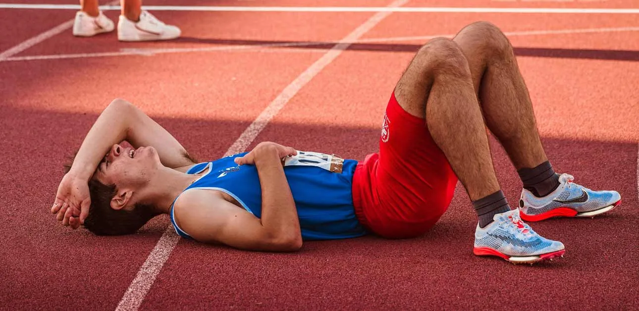 Athlete wearing blue singlet and red shorts, lying down on red athletics track, he's exhausted.